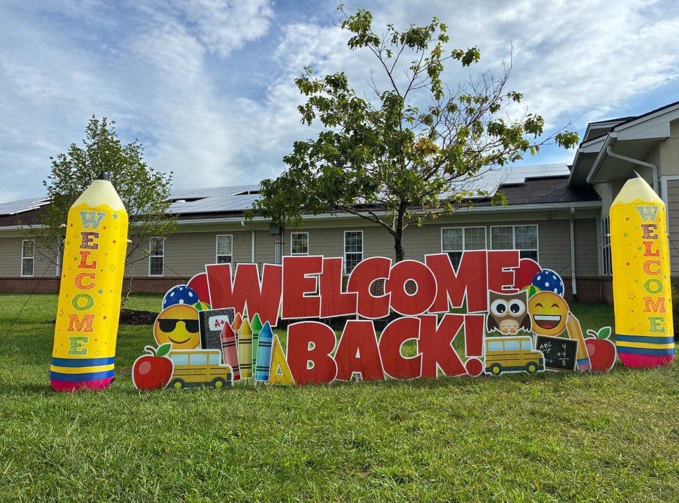 Welcome Back school yard card display with giant pencil inflatables, a school bus, crayons, an owl, and emoji cutouts